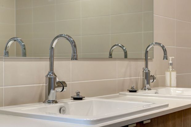 Close-up of a contemporary bathroom vanity with two white ceramic basins, tall polished chrome gooseneck taps, chrome fixtures, beige large-format wall tiles, and a wide mirror reflecting the fittings. Dark timber cabinetry is visible beneath the vanity.