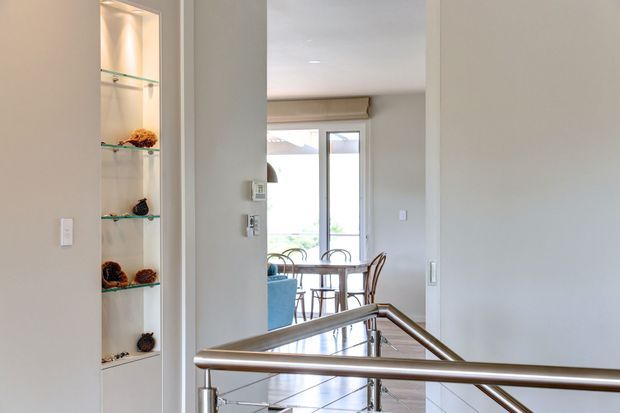 Interior view from a stair landing or hallway looking toward a bright dining area. A stainless-steel handrail and balustrade dominate the foreground, with a recessed illuminated display niche and glass shelves on the left. The dining area beyond has a table, chairs, tiled flooring, and large glazed doors or windows with a roller blind.