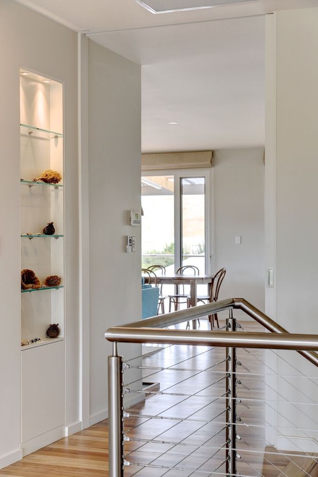 Bright contemporary interior view from a stair landing, looking through an open passage toward a dining area with a glass door and exterior glazing. The foreground features a stainless-steel and cable balustrade, while the left wall has built-in illuminated display shelving. Light timber flooring transitions to pale tiled flooring in the dining area, with neutral painted walls and recessed lighting.