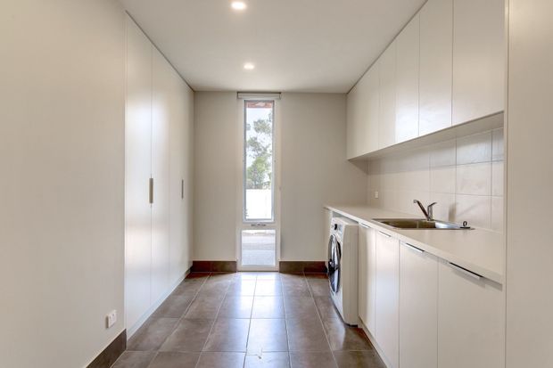 A narrow, compact galley-style laundry and utility space with extensive white built-in cabinetry along both sides. The room has a tiled floor, tiled splashback, stainless-steel sink, washing machine, recessed ceiling lights, and a glazed exterior door at the far end.