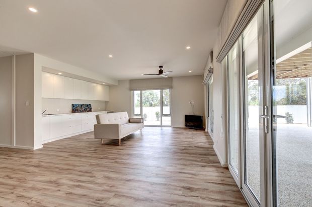 Bright, open-plan living room with warm wood-look flooring, white built-in cabinetry and a compact white sofa. Floor-to-ceiling glazed sliding doors line the right side and open to a covered outdoor area, while recessed ceiling lights and a ceiling fan provide simple contemporary detailing.