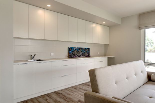 Bright, minimalist open-plan living area with a long wall of white built-in cabinetry, integrated kitchenette, pale tiled splashback, timber-look flooring, and a light upholstered sofa in the foreground. A large window provides natural light, while recessed ceiling lights and a small artwork add subtle detail.