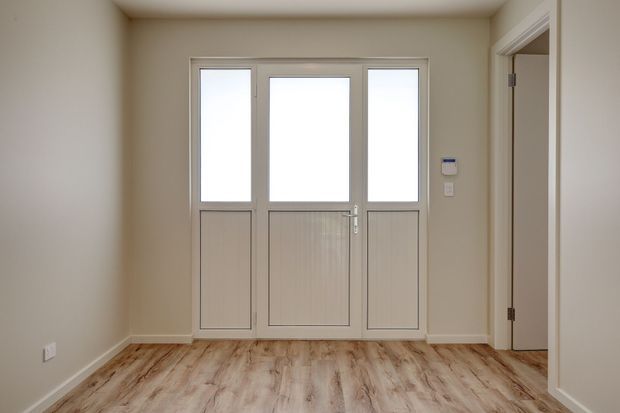 A simple, unfurnished entryway or foyer with a glazed three-panel exterior door, pale neutral walls, white trims, and light wood-look flooring. A doorway opens to another room on the right, while a wall-mounted control panel is visible beside the entrance.