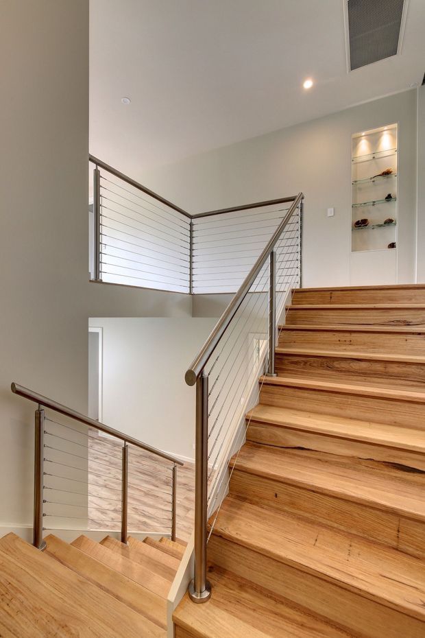 Interior view of a modern multi-level stairway with warm timber treads and landings, slim metal posts, and horizontal cable railings. A recessed illuminated display niche is built into the pale wall beside the upper landing, and the stairwell has a clean open void with recessed ceiling lighting and ventilation.
