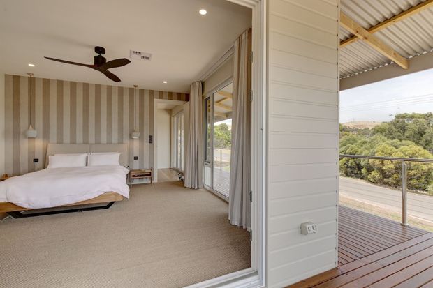 A bright bedroom opening onto a timber deck through wide glazed doors. The room features a low platform bed with white bedding, striped beige-and-cream feature wallpaper, neutral carpeting, full-height curtains, recessed lighting, and a dark ceiling fan. The deck has a corrugated roof supported by exposed timber beams and overlooks a leafy suburban landscape.
