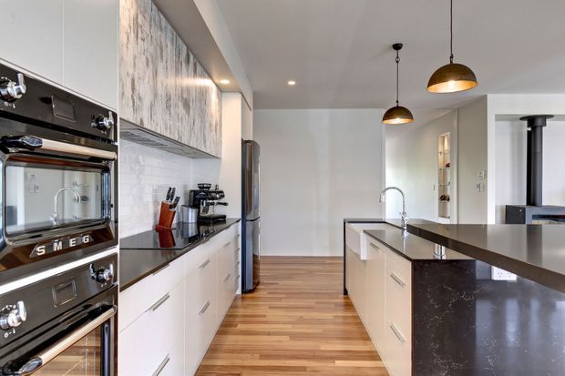 Bright contemporary galley-style kitchen with white flat-panel cabinetry, dark stone benchtops, stainless steel appliances, timber flooring, and two black pendant lights over a central island. A dining or living area is partially visible beyond the kitchen.