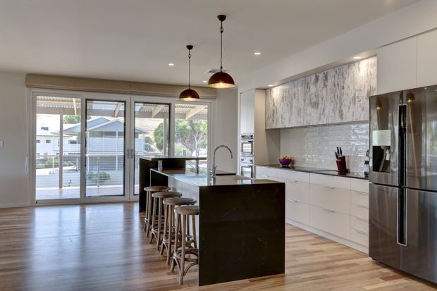A bright, open-plan contemporary kitchen with a large dark island and seating for four, white flat-panel cabinetry, a textured grey-white tiled splashback, and a striking weathered timber-look feature cabinet wall. Stainless-steel appliances, pendant lights, timber flooring, and full-height glazed doors create a spacious connection to the exterior.