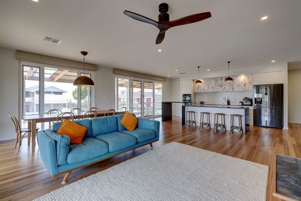 Bright, open-plan contemporary living space with a blue sofa, dining area, and a modern white kitchen with island. The room features warm timber flooring, white cabinetry, pendant lighting, large windows, and a sculptural ceiling fan.