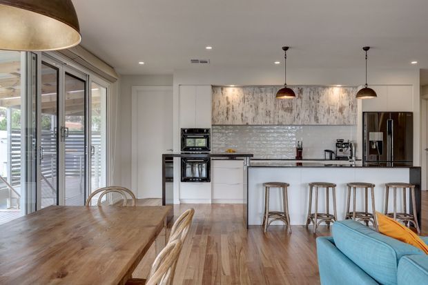 Bright open-plan kitchen and dining area with warm timber flooring and a large timber dining table. The kitchen features white cabinetry, a central island with seating, stainless-steel appliances, a textured light tile splashback, and aged timber-look feature cabinetry. Large glazed doors open to an outdoor area, while pendant lights and a glimpse of a turquoise sofa add contrast.