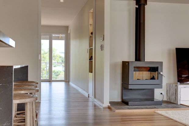 Interior view of a contemporary open-plan living area and hallway. A freestanding wood-burning stove with flue is positioned beside a television and low media cabinetry, while the corridor leads to full-height glazed doors with greenery visible beyond. Timber flooring, pale walls, built-in shelving, and a compact dining or bench area create a warm, uncluttered space.