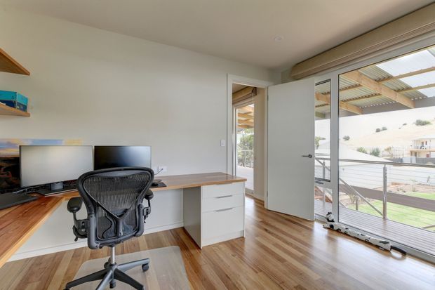 A bright, compact home office with a built-in timber desk and white drawer cabinetry, dual monitors, and an ergonomic black office chair. Full-height glazed doors open to a balcony or exterior walkway, bringing in abundant natural light. Warm timber flooring and a simple white interior create a clean, contemporary workspace.