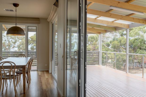 A bright dining area opens through fully glazed folding doors onto a spacious covered timber deck. The interior features warm timber flooring, a pale neutral palette, a large dome pendant light, and a timber dining table with woven chairs. The deck has timber flooring, exposed pale timber beams, a corrugated roof lining, and horizontal metal cable balustrading, with trees visible beyond.