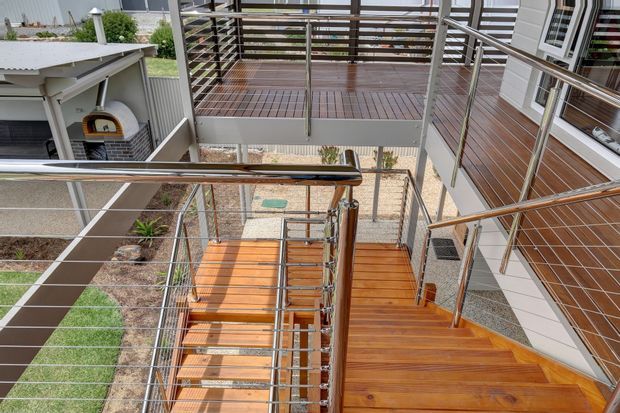 View from an elevated landing down a timber staircase with stainless-steel cable railings, connecting to a raised timber deck and exterior walkway beside a contemporary clad residence. The scene includes a small outdoor courtyard and garden below, with strong horizontal and linear detailing.