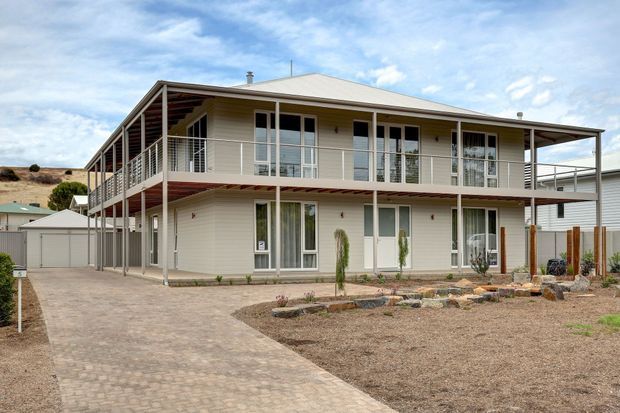 Two-storey residential building with a pale rendered or weatherboard-style exterior, broad wraparound verandah balconies on both levels, simple pitched metal roof, and extensive white-framed glazing. A paved driveway leads past a sparsely landscaped front garden to a detached garage. The architecture has a practical, contemporary coastal or regional character.
