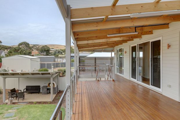 A spacious covered timber deck or veranda running along the exterior of a house, with exposed timber roof beams, a translucent roof, white horizontal cladding, glazed sliding doors, metal balustrades, and timber decking. The deck overlooks a landscaped yard and neighbouring low-rise homes in a hilly suburban setting.
