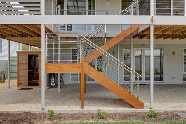 Exterior view of a raised, multi-level residential structure with a prominent timber staircase and stainless-steel cable railings. Light-coloured horizontal weatherboard cladding, exposed timber framing, open undercroft space, and multiple glazed doors and windows are visible. The composition focuses on the external circulation and deck structure.