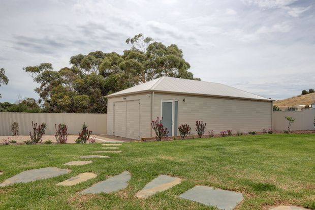 A detached single-storey outbuilding with pale horizontal weatherboard cladding, a light metal hipped roof, a large sectional garage door, and a narrow pedestrian door. It sits in a landscaped grassy yard with stepping-stone paths, low planting, boundary fencing, and mature trees in the background.