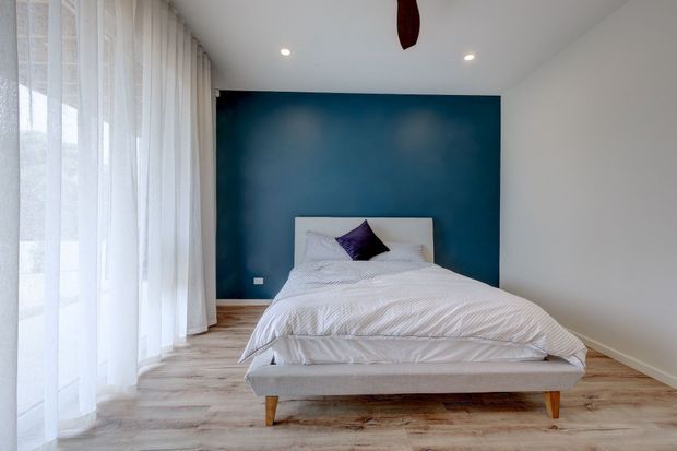 A serene minimalist bedroom with a low-profile upholstered bed centred against a deep blue feature wall. Floor-to-ceiling sheer curtains line the left side, allowing soft daylight into the room, while pale timber flooring and white walls create a bright, calm setting. Recessed ceiling lights and a ceiling fan provide simple functional detailing.