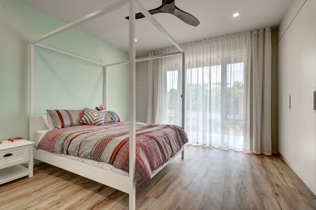 Bright contemporary bedroom with a white four-poster bed, patterned red and neutral bedding, full-height sheer curtains, built-in white wardrobes, and warm wood-look flooring. A ceiling fan and recessed lighting contribute to the clean, airy design.