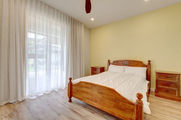 A warmly lit traditional bedroom featuring a substantial polished timber bed with carved detailing and matching bedside furniture. Pale yellow walls, light timber-look flooring, and full-height sheer curtains create a bright, comfortable interior.