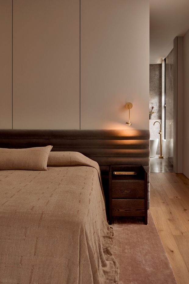 A warm, softly lit bedroom with a large upholstered bed, beige textured bedding, dark brown integrated headboard and bedside storage, and a slim brass wall-mounted reading light. Timber flooring and a muted rug lead toward an open bathroom area with a freestanding bathtub and stone-look wall finish.