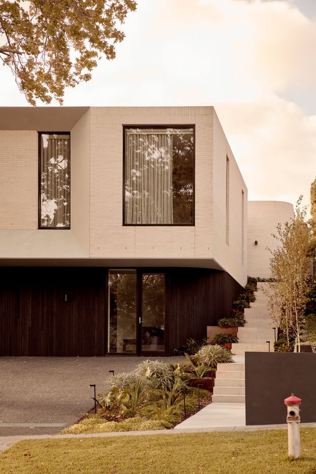 Exterior view of a contemporary two-storey residence with a crisp geometric form, pale textured masonry upper level, dark vertical timber-clad lower level, and large dark-framed windows. A recessed glazed entry sits beneath a projecting upper volume, while a broad exterior stair climbs beside landscaped planting toward the side of the house. The warm-toned image also shows a driveway, lawn, and mature trees.
