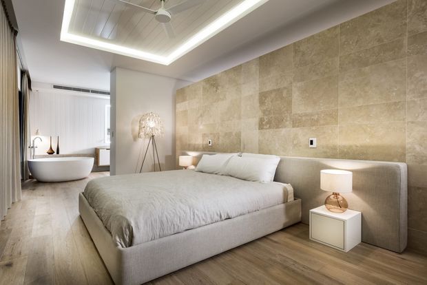 A refined modern bedroom with a large upholstered bed, integrated upholstered headboard, white bedding, pale timber flooring, and a full-height beige stone feature wall. A freestanding white bathtub is visible in an adjoining open bathing area, with soft layered lighting, a sculptural floor lamp, bedside table, and ceiling fan creating a calm, luxurious atmosphere.