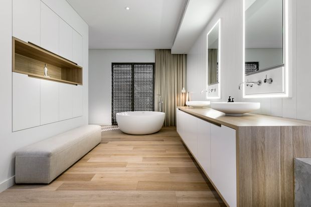 A bright, contemporary bathroom featuring a freestanding white bathtub, a long double vanity with integrated white basins, large illuminated mirrors, and extensive white built-in cabinetry. Warm timber flooring and timber vanity fronts contrast with the white finishes, while a built-in upholstered bench and patterned privacy screens add texture.