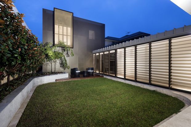 A landscaped courtyard beside a contemporary residential building, featuring a curved lawn edged with concrete paving, dense planting, dark rendered walls, and a long horizontal louvred privacy screen. Large illuminated glazing and concealed outdoor seating create a refined evening setting.
