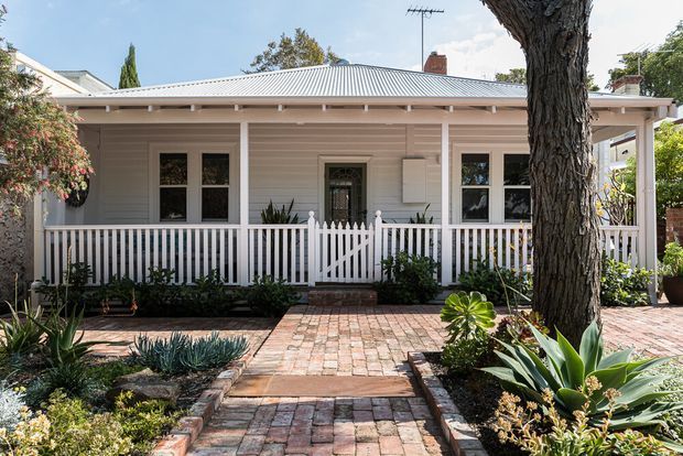 Front exterior view of a single-storey cottage-style home with a pale horizontal weatherboard facade, corrugated metal roof, white trim, and a covered front verandah enclosed by a white picket balustrade. A central gate leads from a brick-paved garden path to the front door, with established low-water landscaping and a mature tree framing the scene.