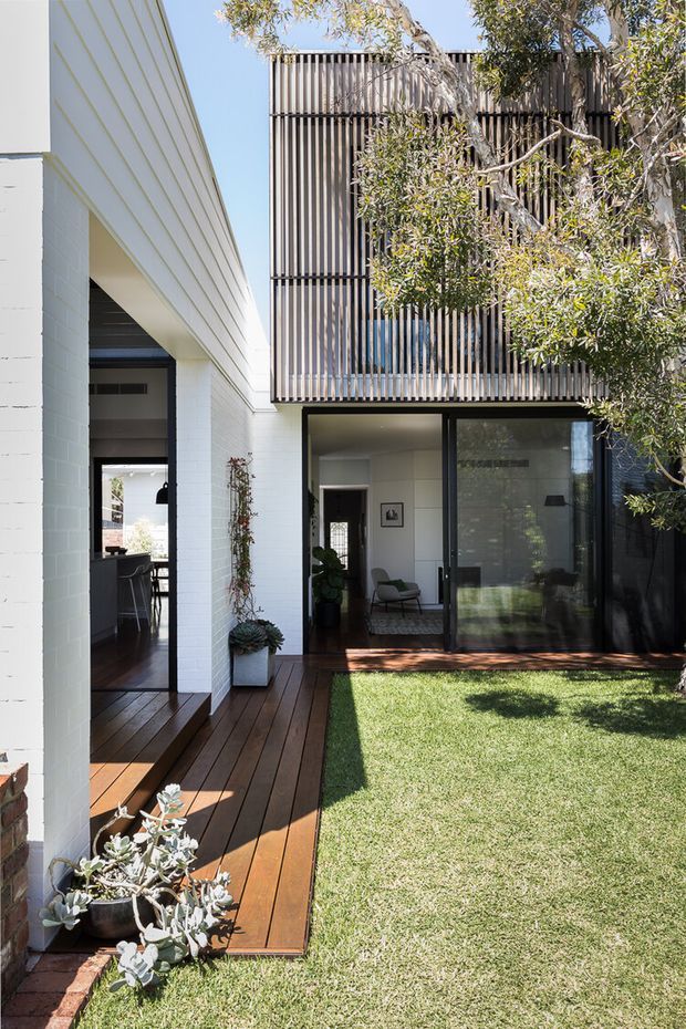 A bright courtyard view of a contemporary multi-level residence. White-painted masonry walls frame a dark timber deck and a small lawn, while large black-framed glazed doors open to an interior living area. The upper storey is screened with closely spaced vertical timber battens, partially obscured by a mature tree. Potted plants and a covered passage add to the landscaped courtyard setting.