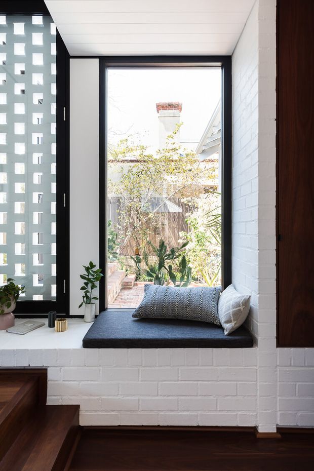 A bright, compact built-in window seat in a residential interior. The seat is set into a white-painted brick wall beneath a large black-framed window, with a dark charcoal cushion and textured grey pillows. A patterned breeze-block screen appears to the left, while dark timber flooring, cabinetry, and stair elements frame the space. Potted plants and a small courtyard are visible through the window. The overall design is clean, warm, and contemporary with strong contrast between white masonry, black frames, and dark timber.