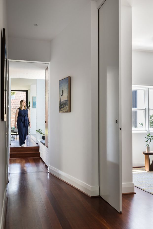 A bright residential hallway with polished dark timber flooring, white walls and trim, and a partially open white door leading into a sunlit room. The corridor includes a framed artwork, recessed ceiling light, and steps toward an adjoining living area with large windows and potted plants. A person is visible in the background.