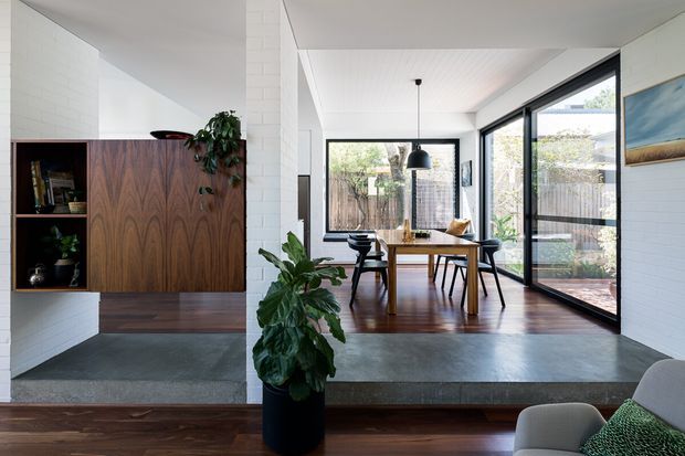 A bright, refined open-plan dining area with a mid-century-inspired timber built-in cabinet, white painted brick walls, polished concrete flooring, and warm dark timber flooring. The dining table sits beside extensive black-framed glazing opening toward a landscaped courtyard. Indoor plants, a sculptural pendant light, and restrained furnishings create a calm connection between interior and exterior.