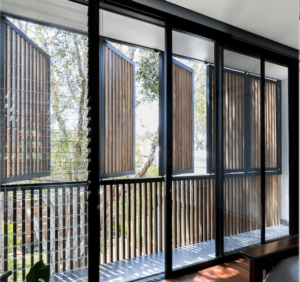 Interior view of a glazed balcony or sunroom enclosure with extensive black-framed floor-to-ceiling glazing, timber vertical privacy screens, and a narrow timber bench. Sunlight filters through the screens, creating striped shadows across the light floor and lower glazing. Trees and a balcony balustrade are visible beyond.