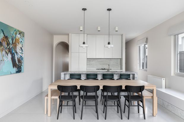 Bright open-plan dining and kitchen interior with a long light timber dining table and black chairs in the foreground. The kitchen has white full-height cabinetry, a pale stone-look splashback and island bench with dark stools. White walls, pale flooring, pendant lighting, large windows and a simple arched doorway create a clean contemporary setting.