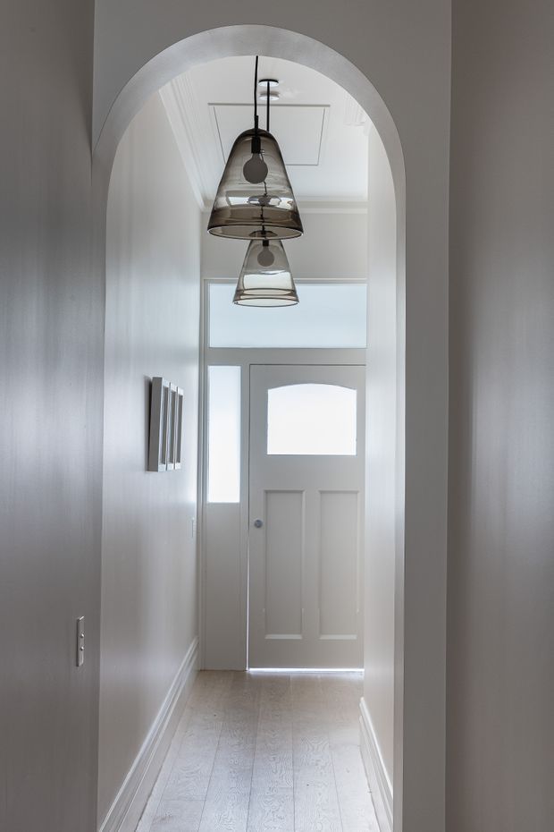 A narrow, bright residential hallway leading to a white entry door with frosted glazing and sidelights. The corridor features a tall arched opening, white decorative ceiling trim, pale timber flooring, grey painted walls, white skirting boards, and two hanging smoked-glass pendant lights.