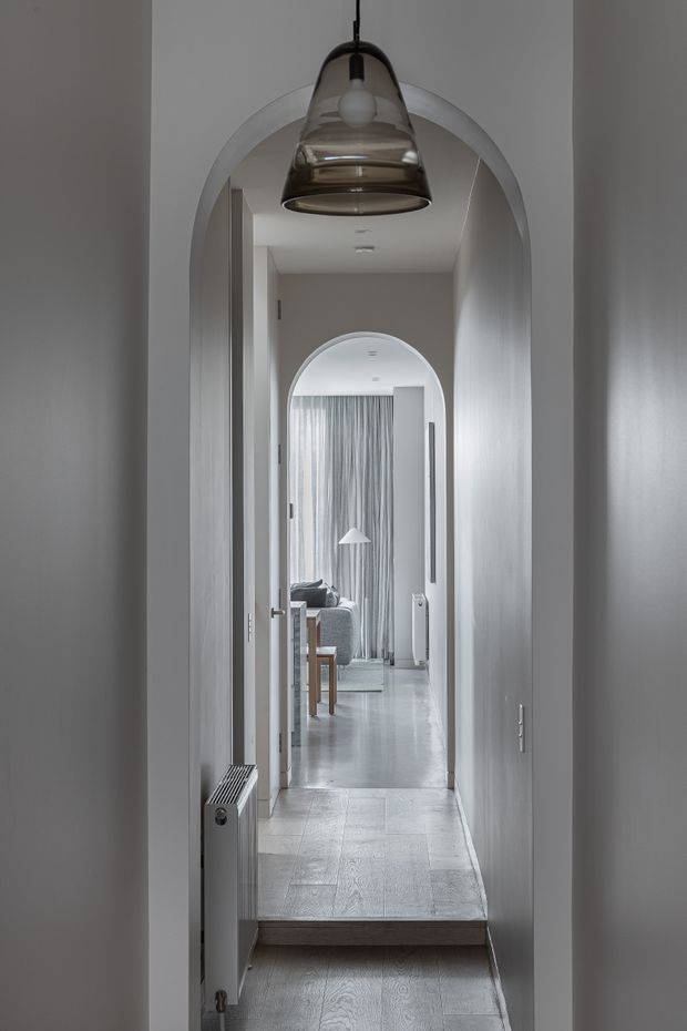 A narrow, softly lit residential hallway with white plaster walls, repeated rounded archways, a tiled or pale timber-look floor, and a dark glass pendant light in the foreground. An open doorway reveals a calm living area with sheer curtains, a sofa, and a floor lamp. A small step separates the hall levels, with a wall-mounted radiator along the left side.