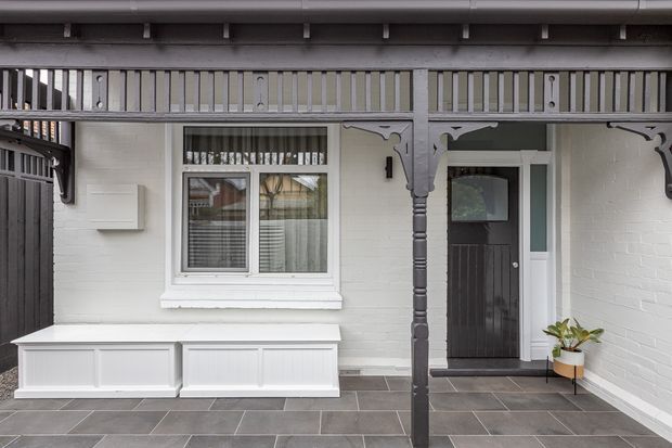 A covered residential entry courtyard or porch with white-painted brick walls, dark grey tiled flooring, a dark timber front door, white-framed window, built-in white storage bench, and ornate timber posts and decorative fretwork supporting the roof. The compact space has a restrained monochrome palette with a small potted plant.