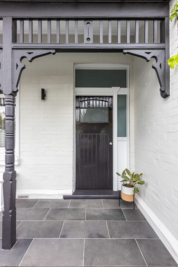 A covered residential entry porch with a dark painted timber door, decorative Victorian-style timber fretwork and posts, white-painted brick walls, and large dark grey tiled flooring. A small potted plant sits beside the entrance.