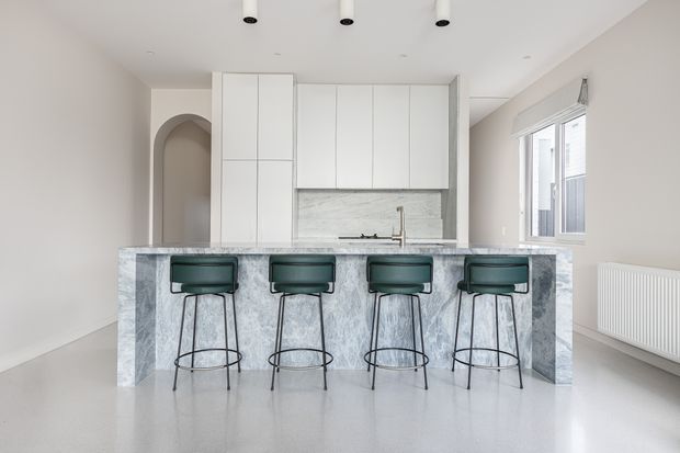 Bright, minimalist kitchen with a large pale marble island and four dark green bar stools. The room features full-height white cabinetry, a matching marble splashback, integrated appliances, light walls and flooring, recessed ceiling lights, an arched doorway, and a large side window with a white radiator beneath it.