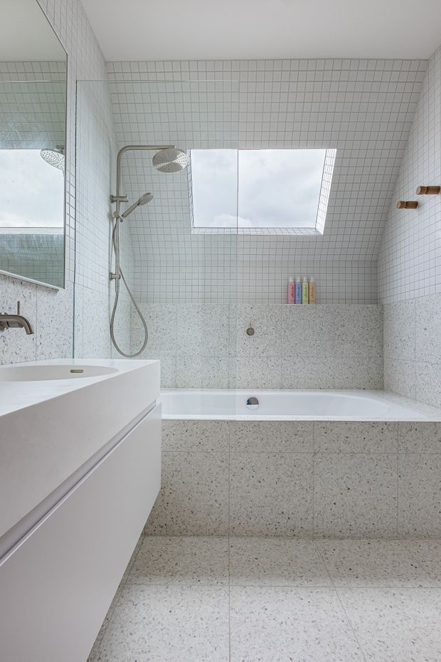 A bright, minimalist bathroom with a white freestanding-style bathtub set into a tiled surround, a wall-mounted white vanity, and a combined rainfall and handheld shower. Small square white wall tiles cover the upper walls, while terrazzo-style grey-and-white tiles clad the lower walls, bath surround, vanity area, and floor. A large skylight brings in natural light, and a mirror reflects the shower area.
