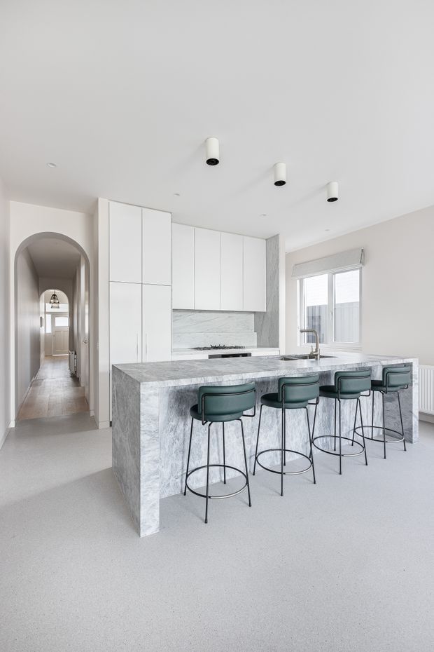 Bright contemporary kitchen with a large grey-veined stone island and waterfall end, white floor-to-ceiling cabinetry, matching stone splashback, integrated cooktop, and undermount sink. Four dark green upholstered bar stools line the island. The space connects to a hallway through an arched opening and has large windows, a pale floor, and simple cylindrical ceiling downlights.