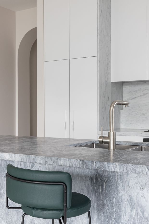 A refined contemporary kitchen viewed from the island, featuring extensive pale grey marble or stone surfaces on the island benchtop, waterfall edge and splashback. Integrated white cabinetry provides tall storage and overhead cupboards, while a brushed metal gooseneck tap and undermount sink sit within the island. A dark green upholstered bar chair adds contrast, and a rounded archway is visible in the background.