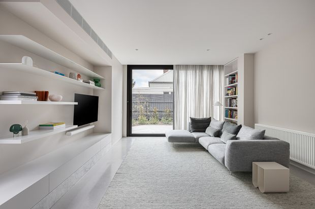 Bright contemporary living room with a large grey sectional sofa, pale textured rug, built-in white media and display joinery, integrated television, open shelving, full-height sheer curtains, and a large glazed door opening to a small exterior courtyard. A radiator and bookshelf are visible along the right wall.