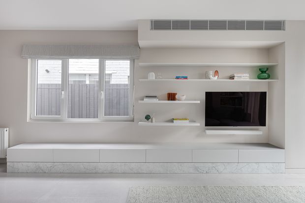 Minimalist contemporary living room media wall in a pale neutral palette, featuring a long built-in white storage unit with a marble-look base, recessed open shelving, and a wall-mounted television. A large window with a roller blind provides natural light, while a linear air-conditioning vent is integrated above the joinery.