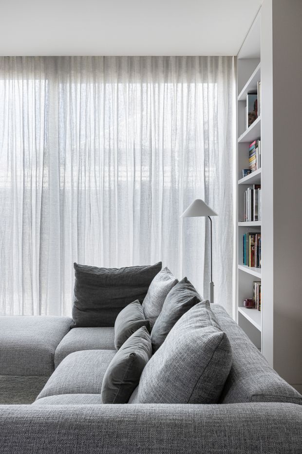 A softly lit, contemporary living room corner dominated by a large grey sectional sofa with layered grey cushions. Floor-to-ceiling sheer curtains diffuse daylight across the room, while a slim grey floor lamp and white built-in open shelving filled with books provide functional details. The palette is restrained in white and grey tones with a calm, minimal appearance.