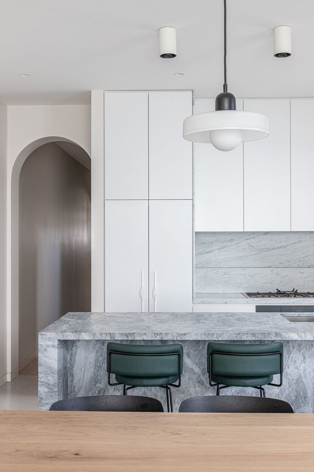 A refined contemporary kitchen viewed from the dining area, featuring a large grey-veined stone island with integrated seating, white full-height cabinetry, a matching stone splashback, dark green upholstered bar stools, and a sculptural pendant light. A tall arched doorway opens to a darker adjoining space, while a timber dining table occupies the foreground.