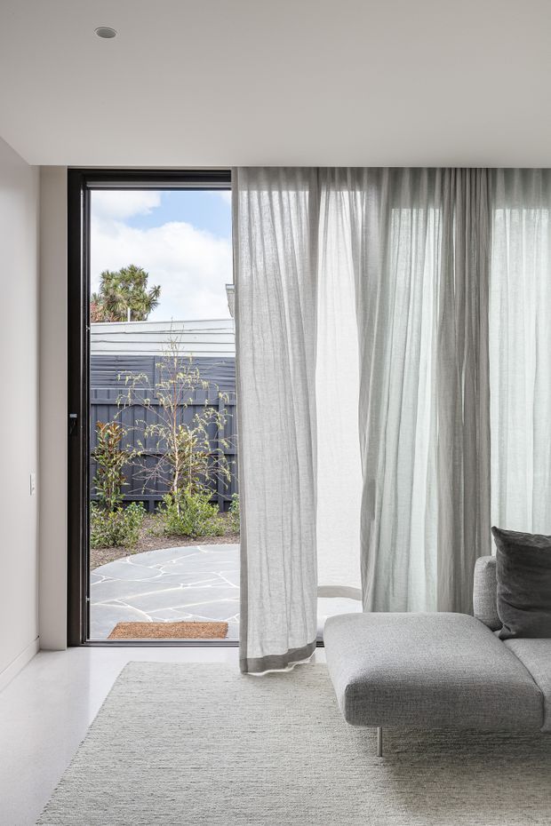 A minimalist, light-filled living room corner with a large dark-framed glazed door opening to a small paved courtyard. Sheer floor-to-ceiling curtains, a low grey sectional sofa, and a pale textured rug create a soft neutral interior. The room has a smooth white ceiling and walls, with a view of dark fencing and planting outside.