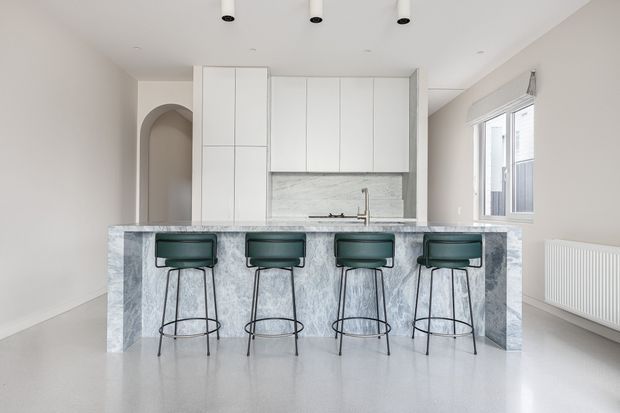 Bright minimalist kitchen with a large marble-look island and four dark green bar stools. White floor-to-ceiling cabinetry, a pale stone splashback, white walls, an arched doorway, recessed ceiling lights, and a large window create a clean, airy interior.